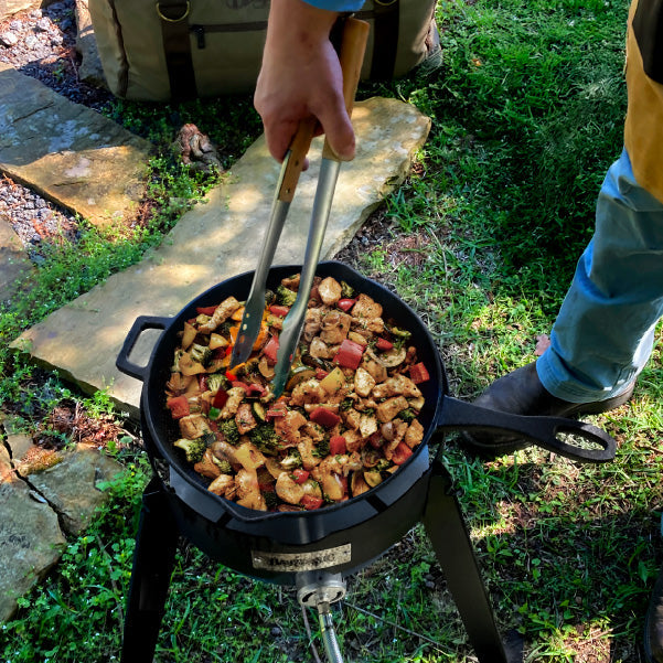 Man stirring chicken and vegetable is a cast iron skillet on a burner outside