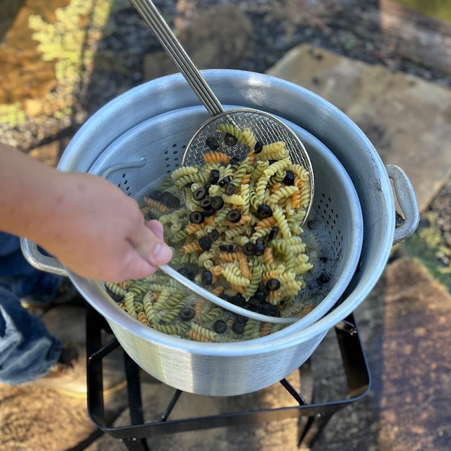 Pasta being drained in an aluminum basket over a pot outdoors