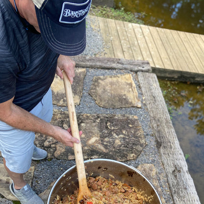 Person cooking outdoors by a body of water, stirring food in a stainless pot with a wooden spoon.