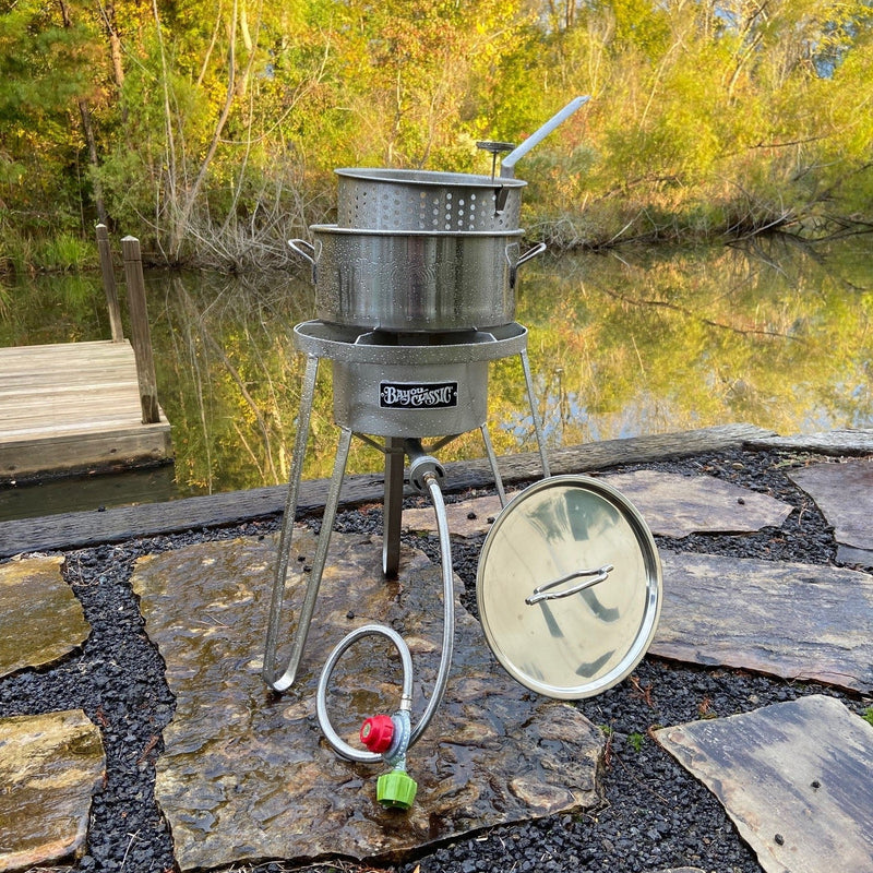 Outdoor stainless cooker with stainless fry pot sitting on a patio setting by water with autumn trees in the background.