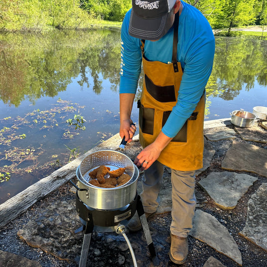Person cooking outdoors by a lake using a fry pot and outdoor cooker.