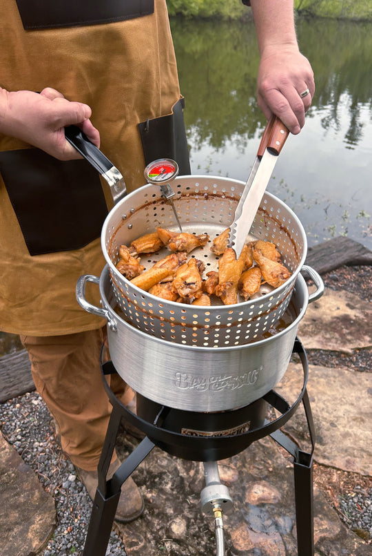 Person cooking wings in a metal pot over a fire outdoors by a body of water.