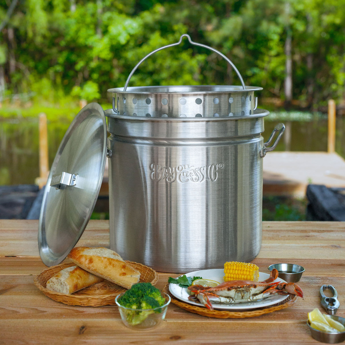 A large aluminum pot with a lid and steamer insert sits on a wooden table outdoors, surrounded by food items like bread, broccoli, crab, and corn, with a blurred green forest background.