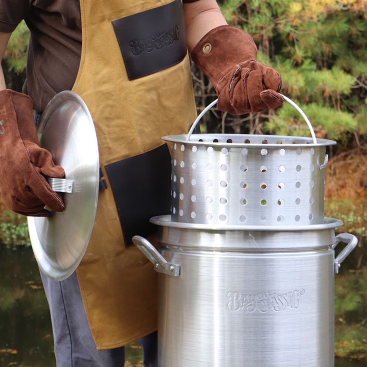 Person outside wearing an apron and brown fry gloves lifting a basket from a stockpot