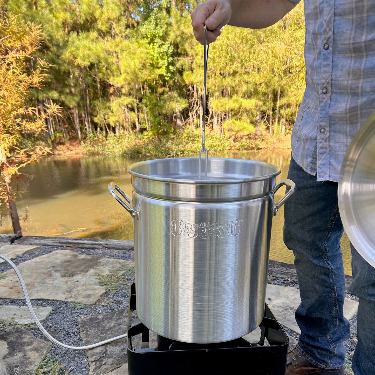 Person using a 42-qt aluminum turkey fryer and outdoor cooker near a body of water.
