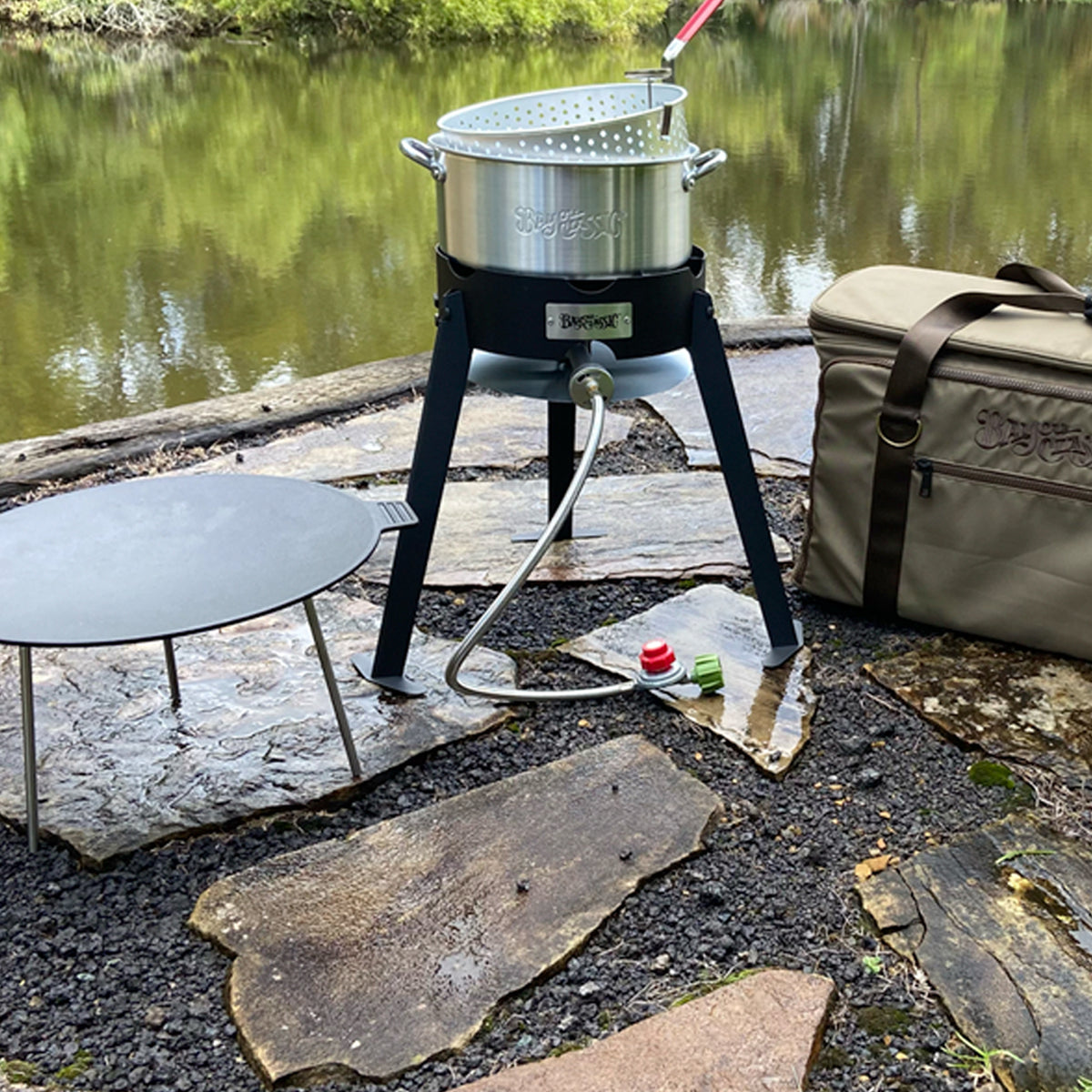 Tall steel cooker with aluminum fry pot, cast iron discada, and gear bag by a body of water