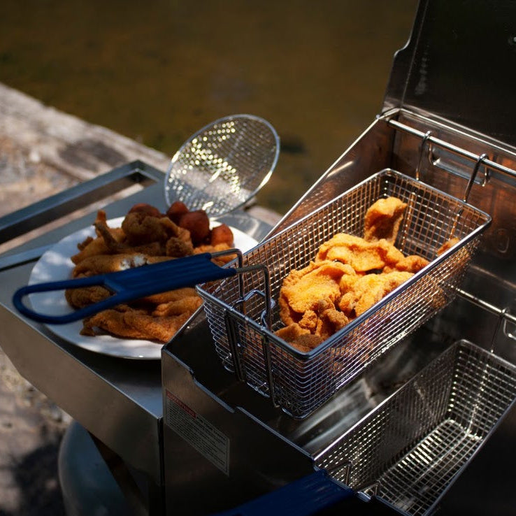 A stainless steel deep fryer with a basket filled with golden fried food, shown outdoors beside a stone surface and water. A plate with more fried pieces and a blue-handled strainer sit on the side.