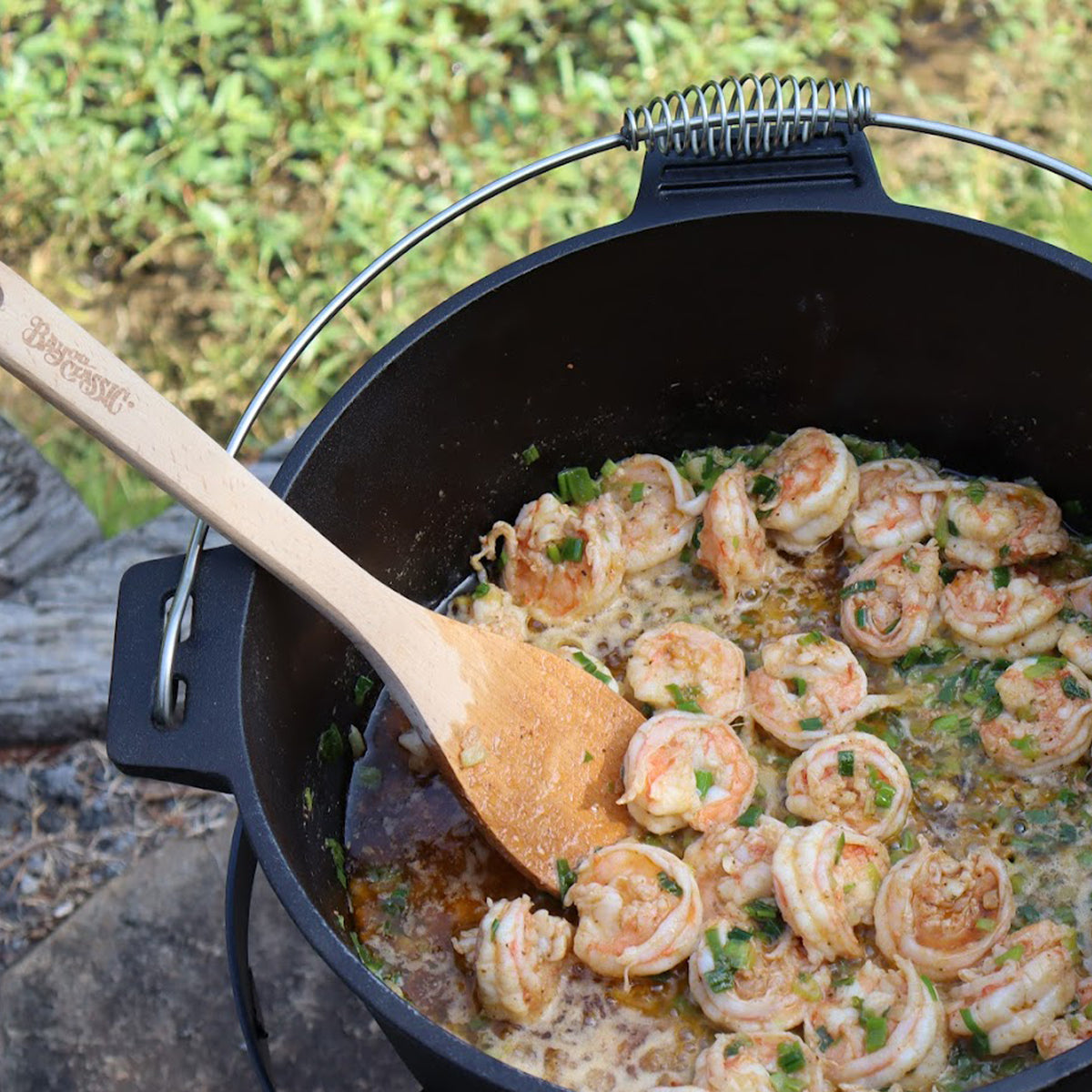 Shrimp cooking in a black cast iron Dutch oven with a wooden spoon, outdoors.