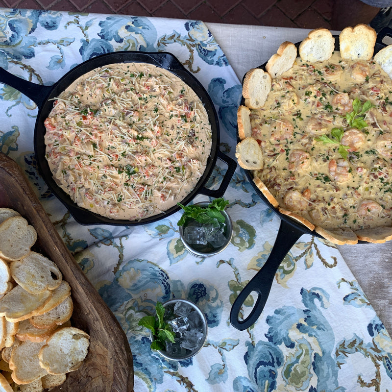 Two cast iron skillets of dip with bread slices on a patterned tablecloth.