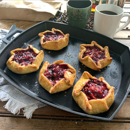 Pastry tarts with berry filling on a black pan with coffee and tea cups in the background.