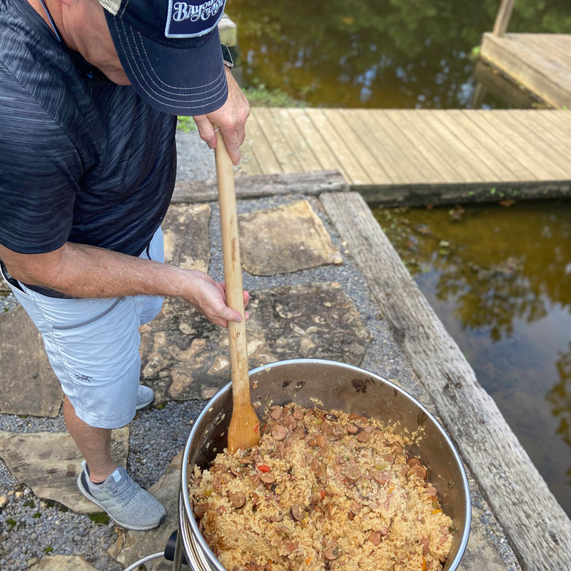 Man cooking jambalaya outside using a stainless kettle and wooden stir paddle by a body of water