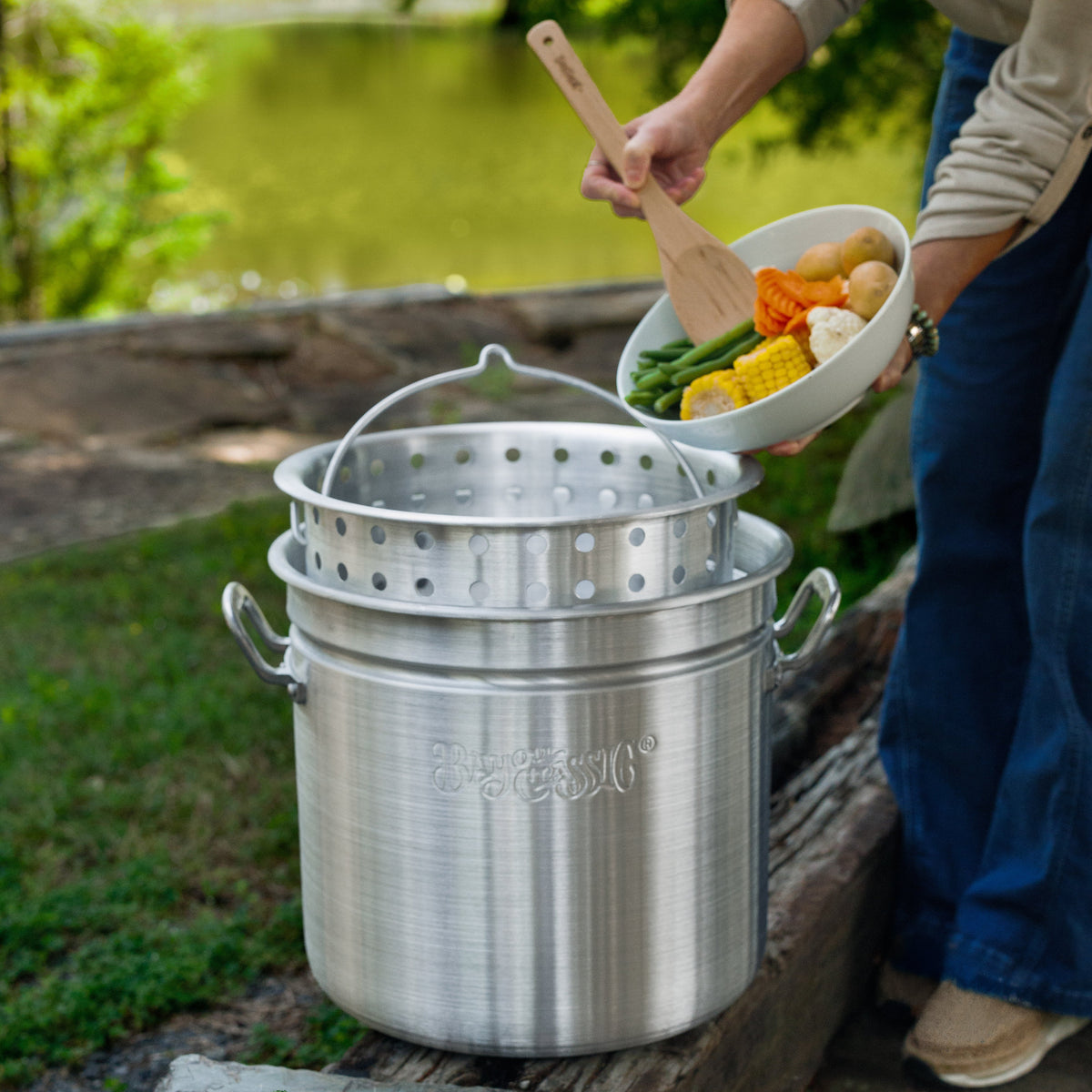 Person holding a bowl of vegetables near a large aluminum pot outside by a body of water.