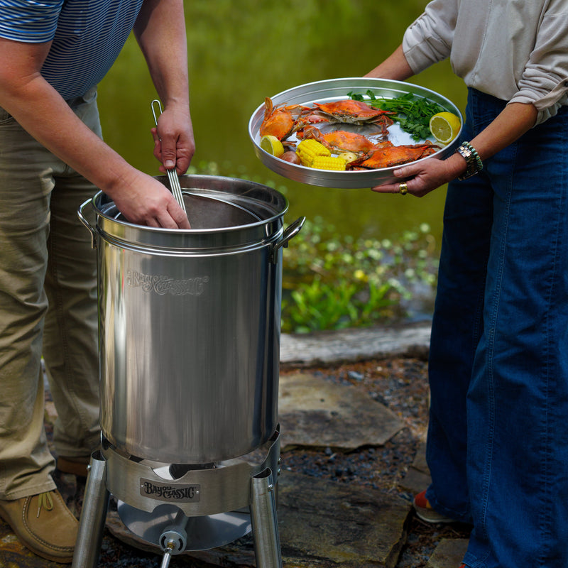 A stainless steel outdoor boiling pot and cooker setup with two people in a natural setting. One person serving crabs on a tray with lemons and seasoning in an outdoor setting.