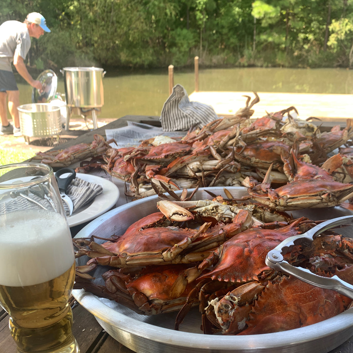 Cooked crabs on a table with person using a stainless cooker kit in the background by a body of water