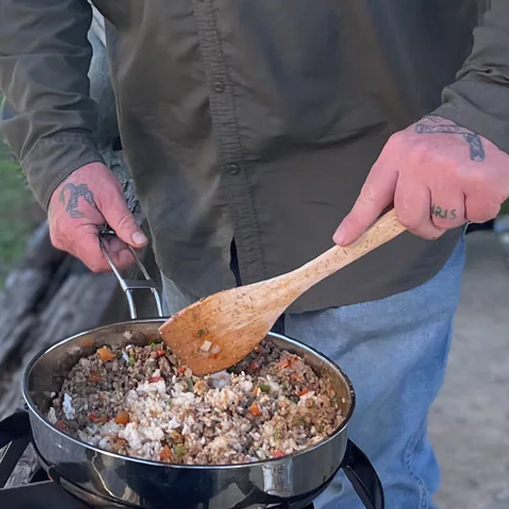 Person cooking outdoors with a stainless pan of food and wooden spoon
