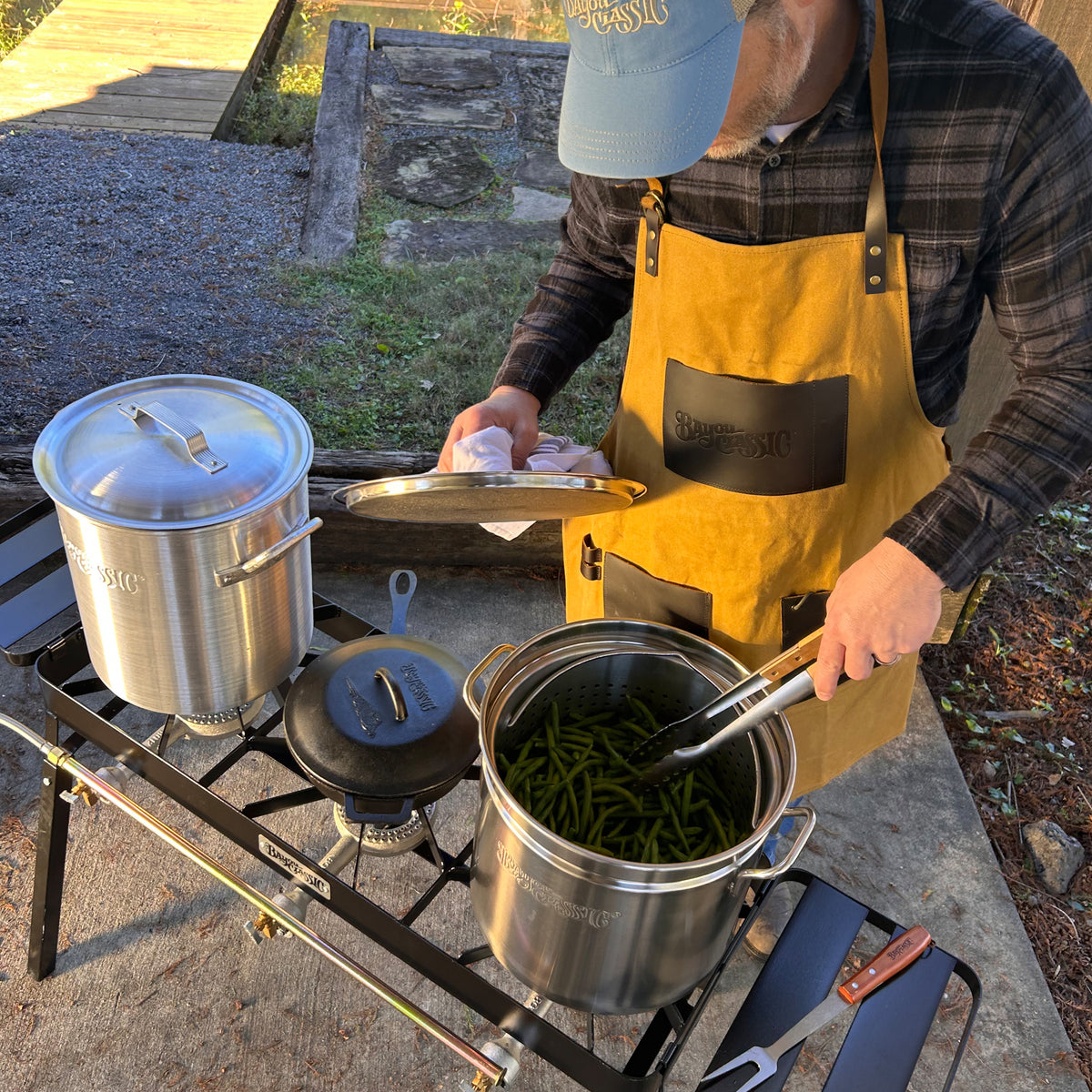 Person outside using triple burner cooker with stainless pot of green beans, an aluminum pot, and a cast iron skillet