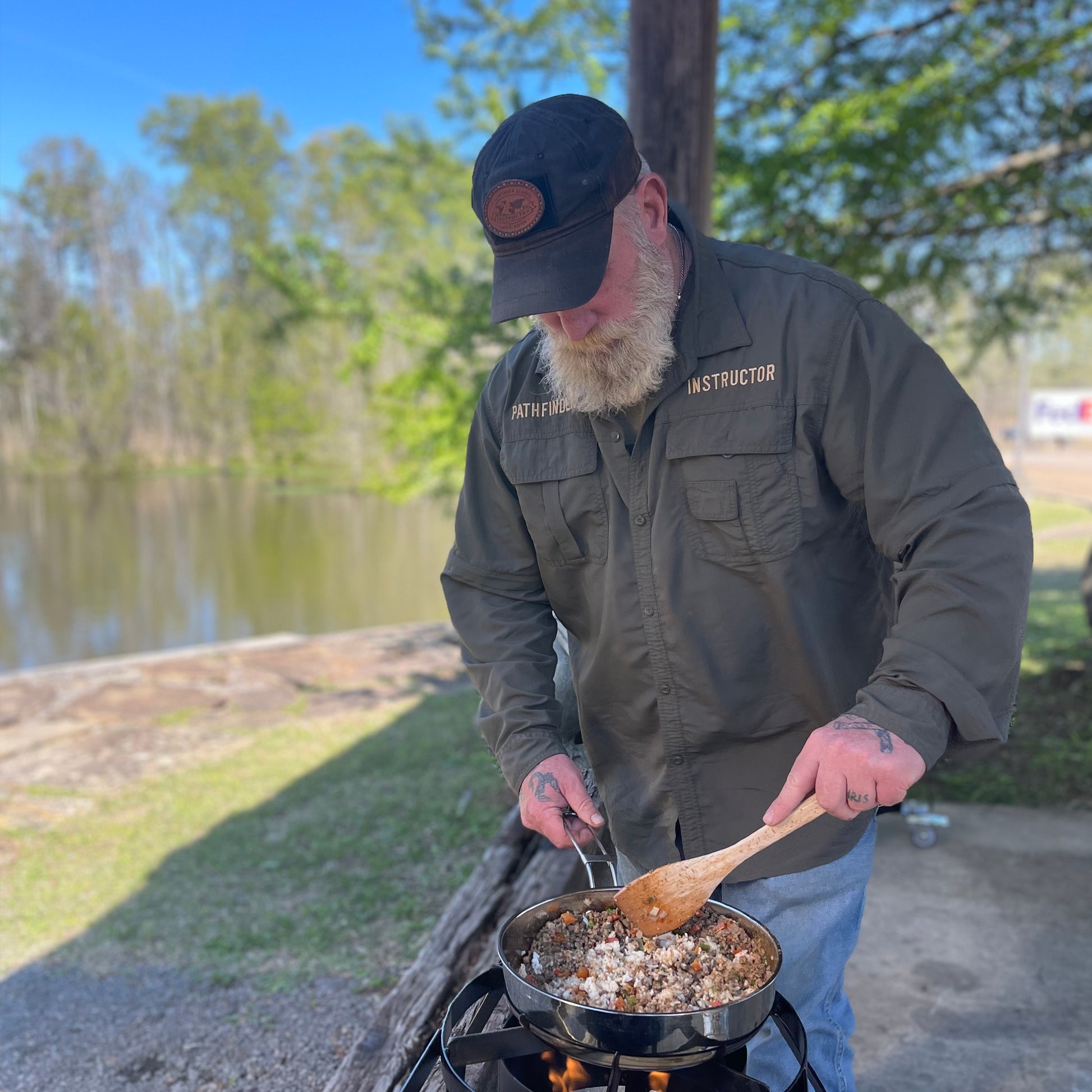 Man cooking outdoors with a stainless fry pot and outdoor cooker by a lake, wearing an instructor shirt and cap.