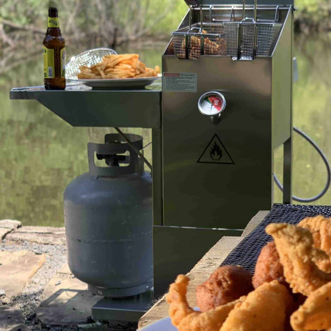 stainless outdoor fryer with fried food on a side shelf. fried fish on a table by a body of water.