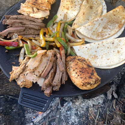Cooked strips of steak and chicken with grilled peppers, onions, and fajita wraps on a cast iron discada over a campfire.