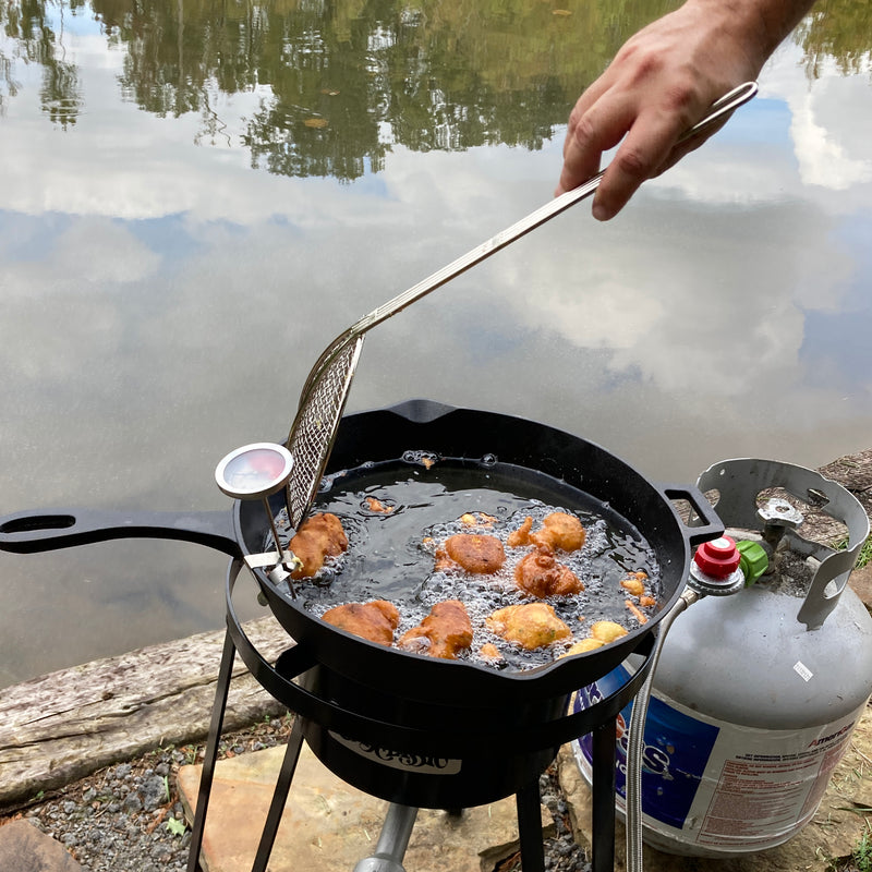 Person frying hushpuppies in a cast iron skillet over a cooker outside 