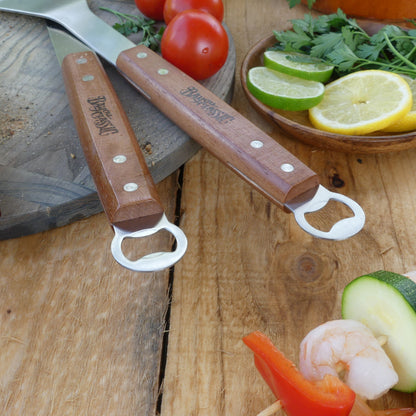 The bottle opener end of the spatula and tongs on a wood surface with vegetables.